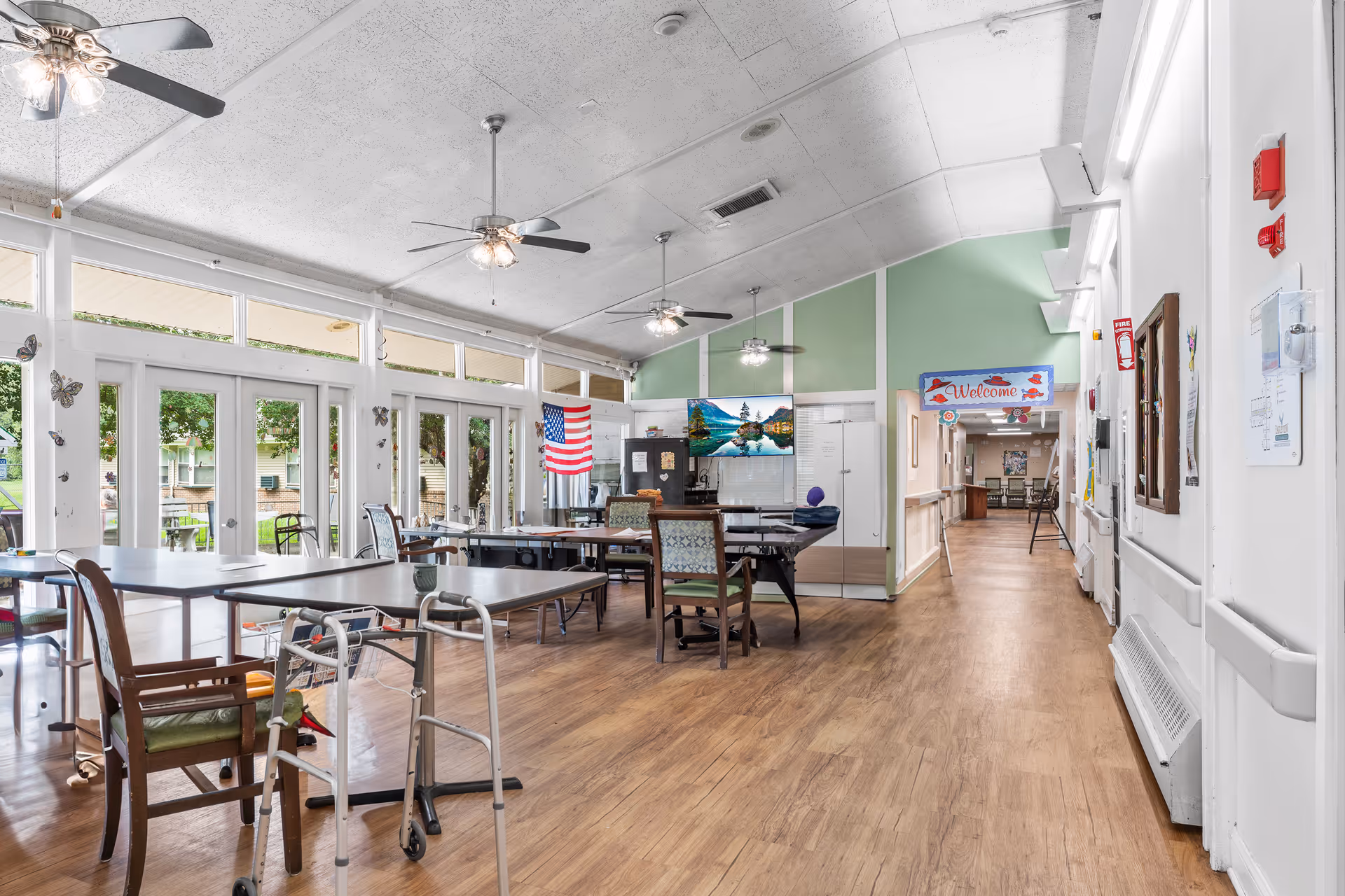 A bright and spacious common area in a senior living facility with multiple tables and chairs arranged for group activities or dining. The room features large windows and glass doors letting in natural light, ceiling fans, an American flag, and a wall-mounted TV. A hallway with a 'Welcome' sign is visible in the background.