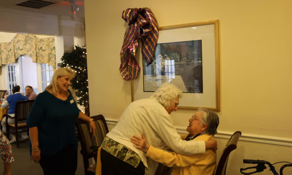 Two elderly women warmly embracing each other in a common area of a senior living facility, while a staff member smiles nearby. In the background, other residents are seated at tables, and a decorated plant with lights is visible.