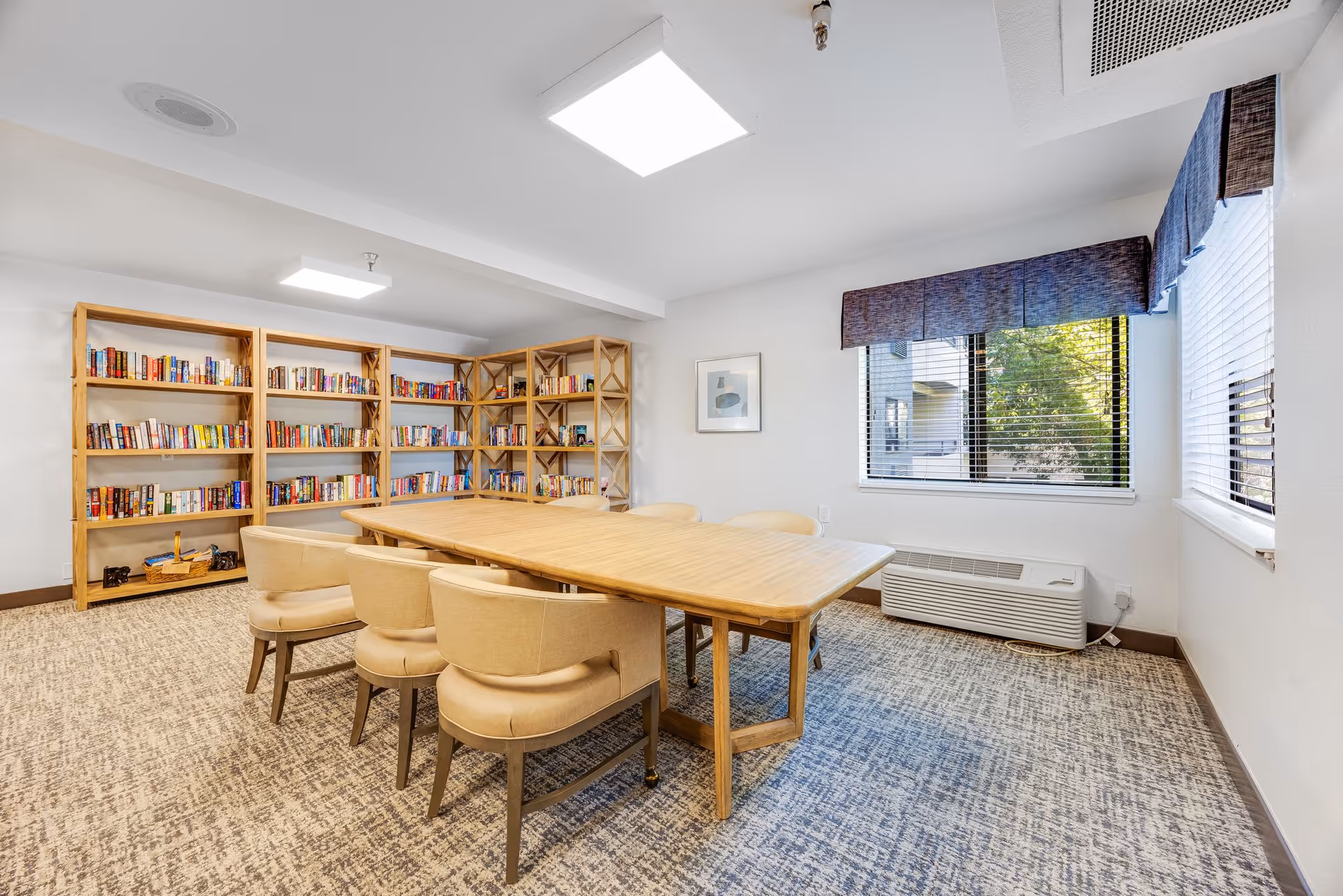 A bright room with a large wooden table surrounded by six beige upholstered chairs. Along the back wall are wooden bookshelves filled with books. Two windows with blue valances let in natural light, and a framed picture hangs on the wall. The floor is carpeted in a patterned design.