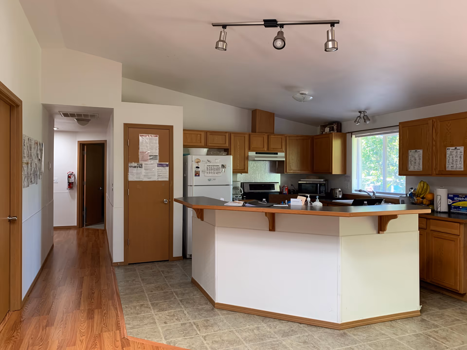 Interior view of a kitchen area in Comfort Haven Adult Family Home featuring wooden cabinets, a white refrigerator, a stove with a range hood, a microwave, and a kitchen island with a wooden countertop. The floor is tiled in the kitchen area and transitions to wood flooring in the adjacent hallway. There is a window letting in natural light and various kitchen items on the counters.