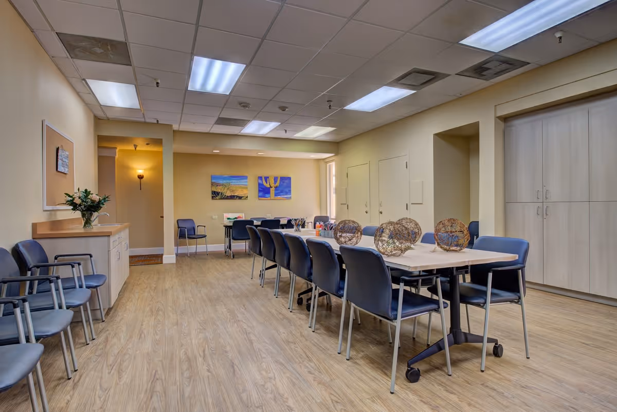 A spacious room with a long table surrounded by blue chairs, decorated with spherical wicker centerpieces. The room has light wood flooring, beige walls, and a ceiling with fluorescent lights. There are additional chairs lined up against the left wall, a small cabinet with a vase of flowers, and two colorful paintings on the far wall.