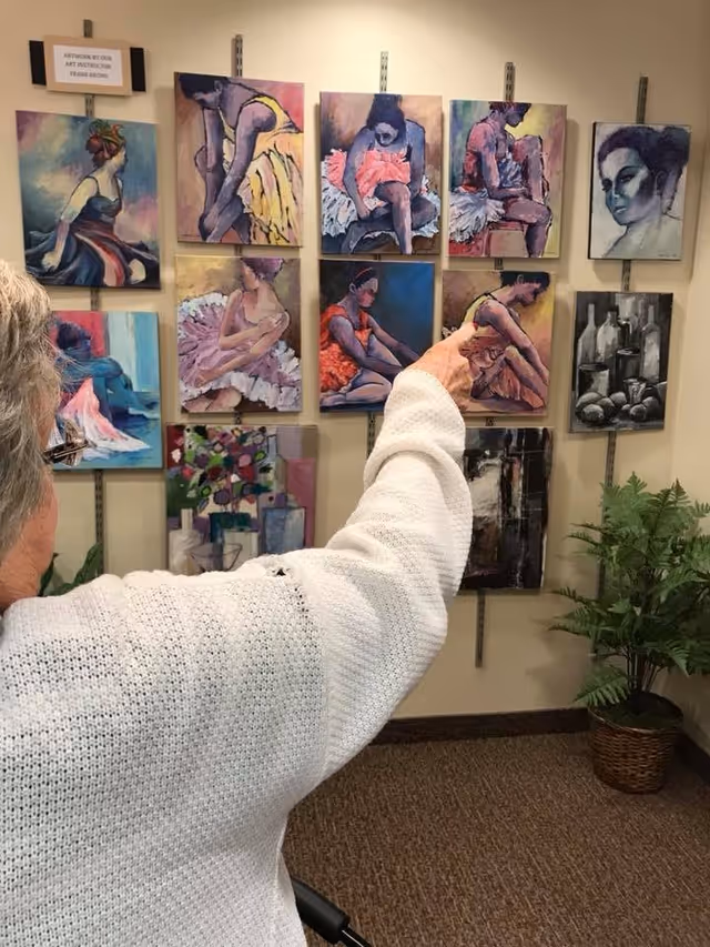 An elderly person wearing a white sweater is pointing at a wall displaying multiple colorful paintings, mostly of ballerinas in various poses. There is a small sign above the paintings that reads 'ARTWORK BY OUR ART INSTRUCTOR FRANK BROWN'. A potted plant is visible on the floor to the right.