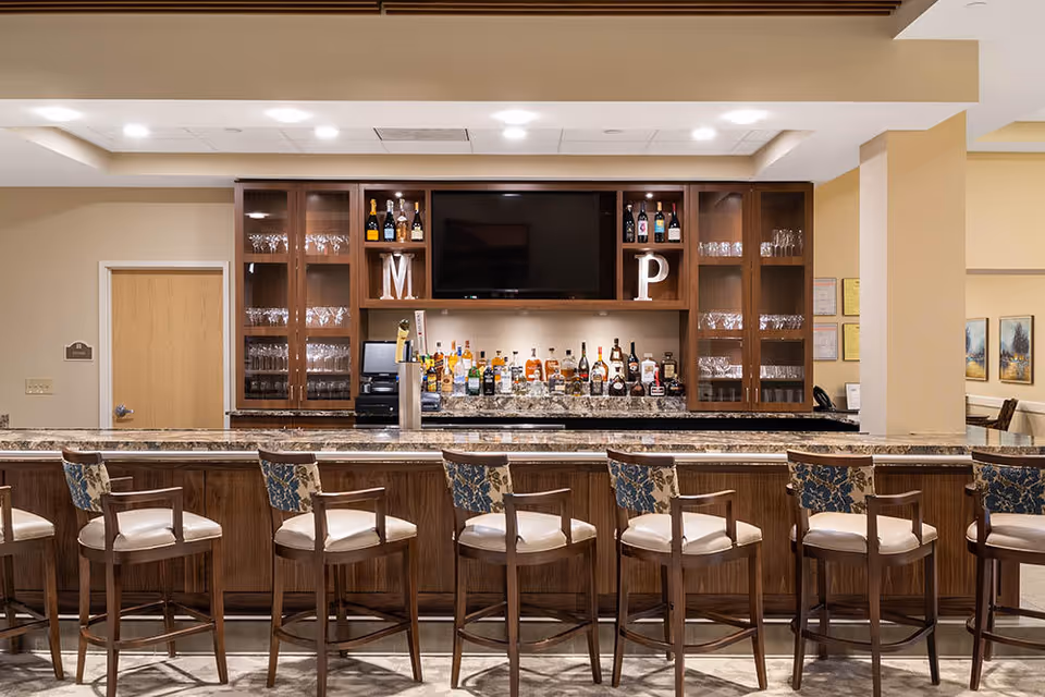 Well-lit indoor bar area with a long marble-topped counter, a row of bar stools, liquor bottles on the back shelf and glass-front cabinets under a mounted TV.