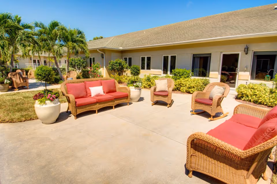 Outdoor patio area at Lynmore at Lawnwood featuring wicker furniture with red cushions, including two sofas and three chairs, surrounded by potted plants and greenery under a clear blue sky.