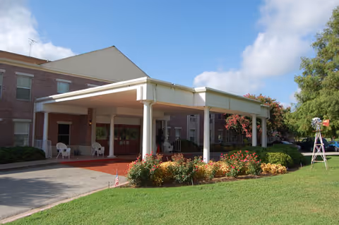 Covered entrance canopy of a brick senior living building with white columns, outdoor chairs, landscaped flowerbeds, and a green lawn under a blue sky.