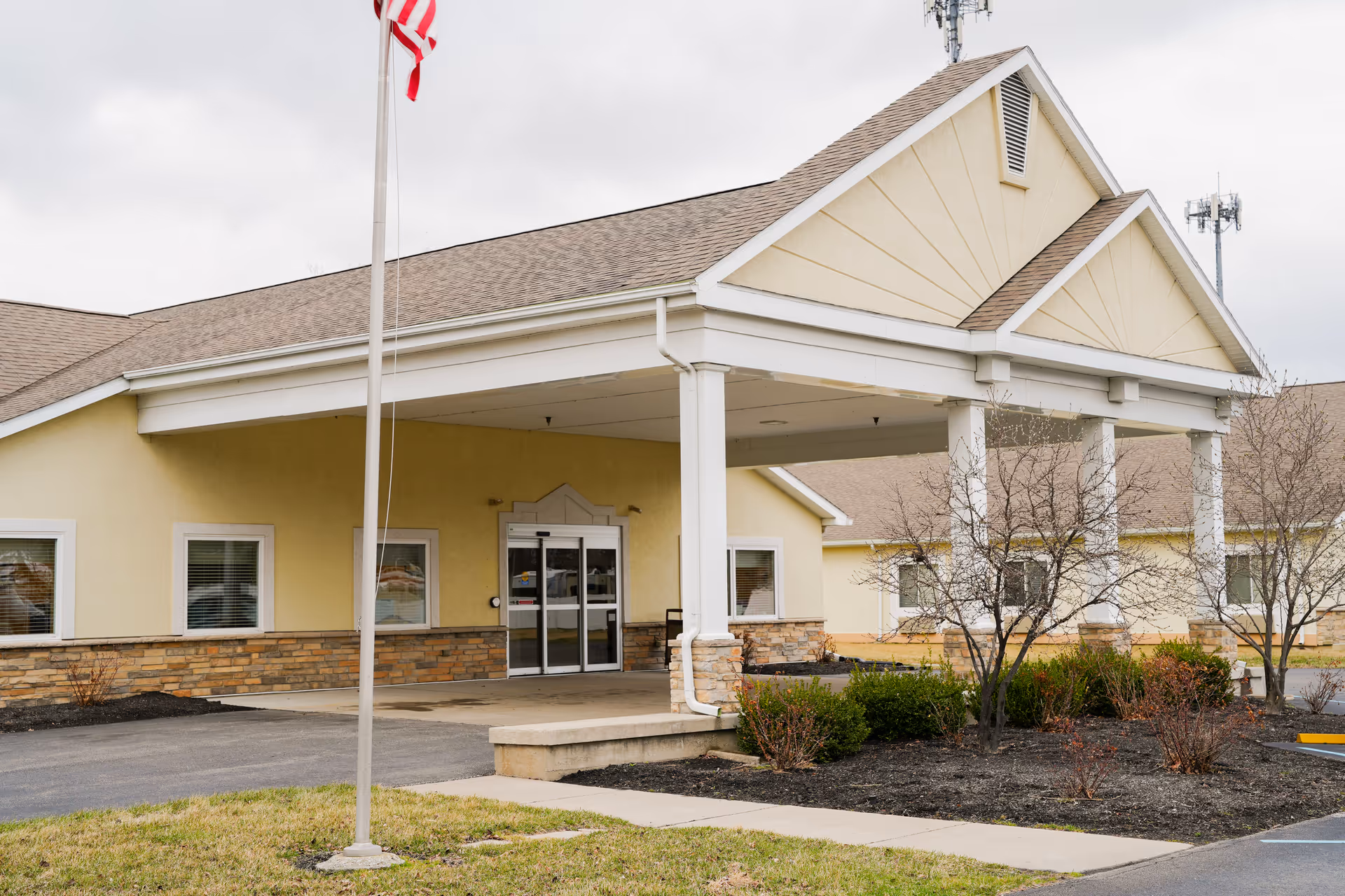 Exterior view of Majestic Care of Point Place building entrance with a covered drop-off area, beige walls, stone accents, and an American flag on a flagpole in front. There are small bushes and leafless trees in the landscaped area near the entrance.