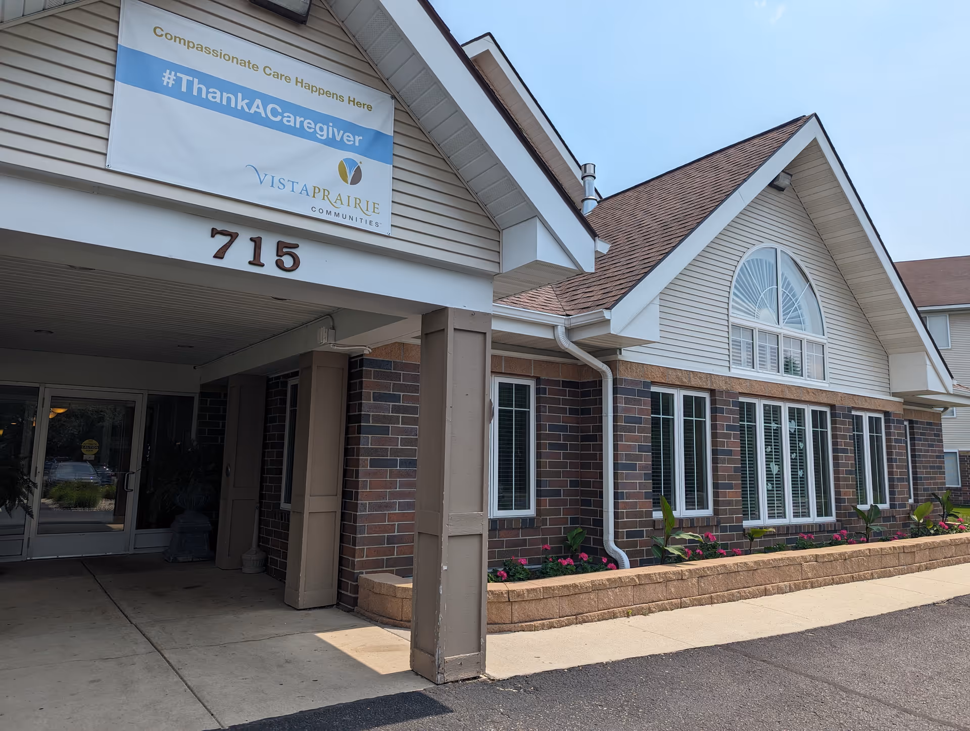 Exterior front entrance of Vista Prairie at Windmill Ponds facility showing a covered walkway with the address number 715. A banner above the entrance reads 'Compassionate Care Happens Here #ThankACaregiver' with the Vista Prairie Communities logo. The building features brick and siding with multiple windows and a flower bed along the front.