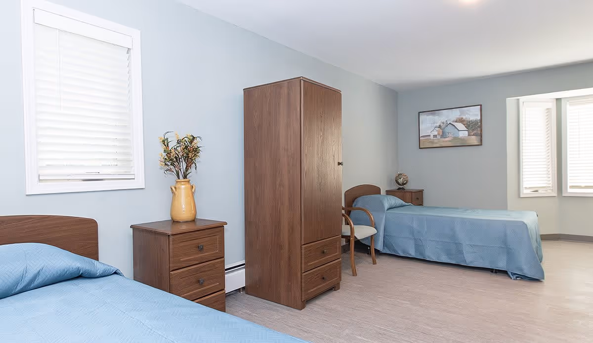 A simple bedroom with two single beds covered in blue bedspreads, a wooden wardrobe, a small wooden nightstand with a yellow vase holding flowers, a wooden chair with a cushion, and a framed picture on the wall. The room has light blue walls and a window with white blinds.