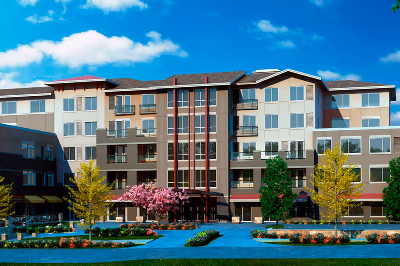 Front exterior view of a multi-story senior living facility building with large windows, balconies, and a landscaped area featuring trees and flower beds under a blue sky with some clouds.