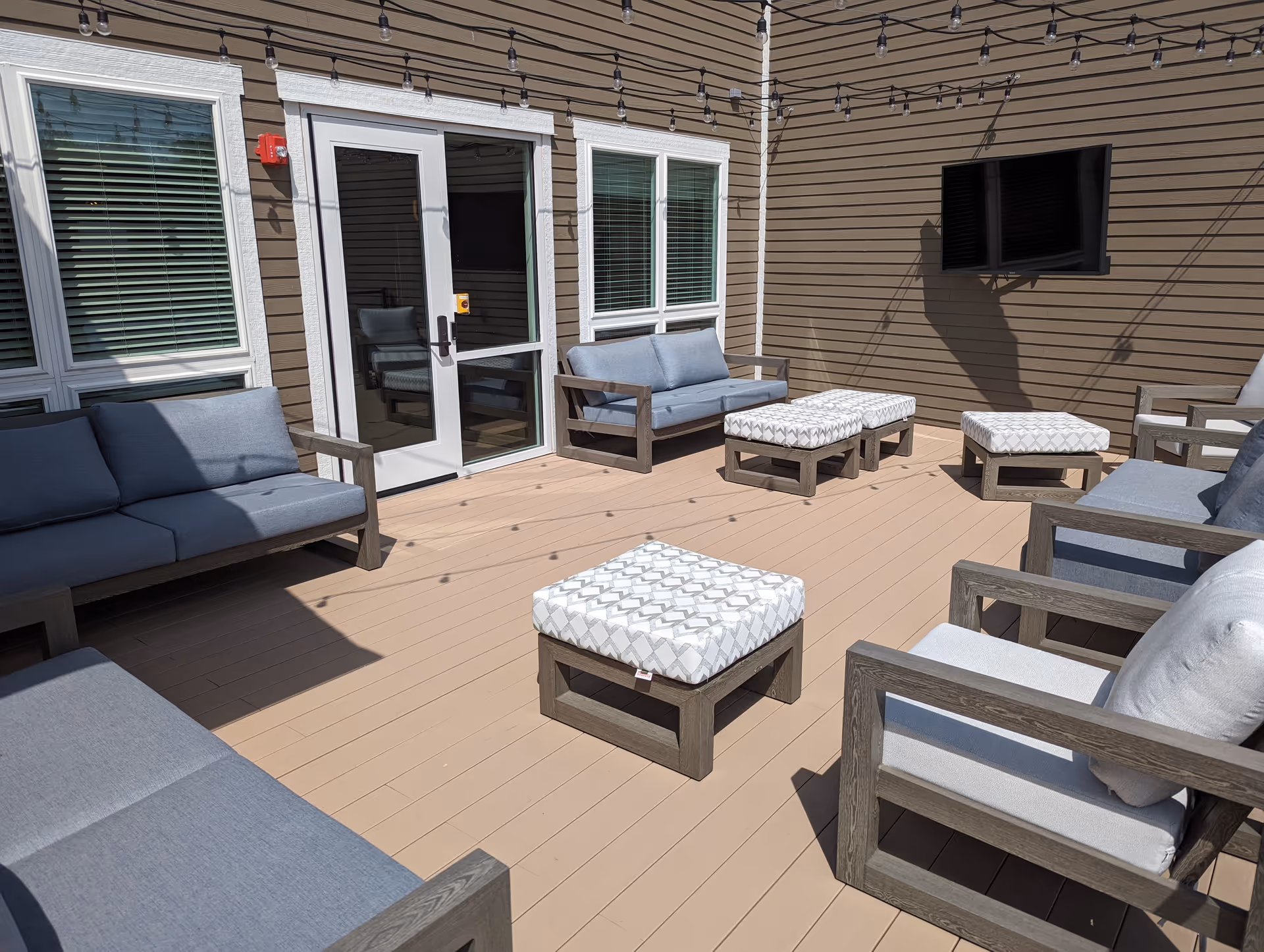 Outdoor patio area with beige wooden flooring, several cushioned chairs and sofas with gray cushions, patterned ottomans, string lights overhead, and a wall-mounted flat screen TV on the brown siding exterior of the building.