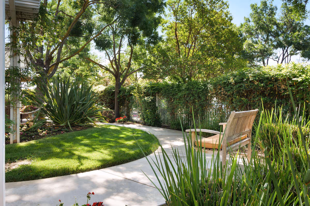A peaceful outdoor garden area with a curved concrete pathway, green grass, various plants, trees providing shade, and a wooden bench with a cushion along the path.