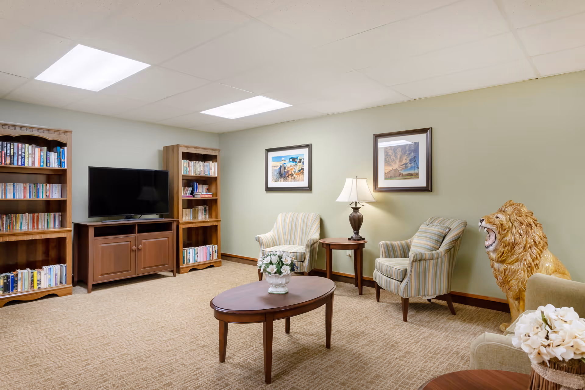 A cozy living room with beige carpet, two striped armchairs, a wooden coffee table with a flower vase, a side table with a lamp, two wooden bookshelves filled with books, a flat-screen TV on a wooden stand, and a large decorative lion statue near the wall. Two framed pictures hang on the light green wall.