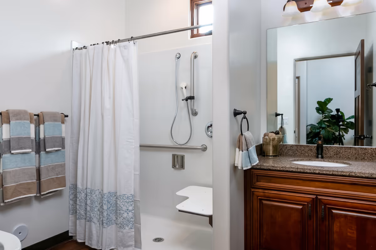 A bathroom featuring a walk-in shower with a white curtain, grab bars, and a fold-down shower seat. Next to the shower is a wooden vanity with a granite countertop, an oval sink, and a large mirror above. Towels in shades of brown, blue, and white hang on a towel rack and a ring. A small window is visible above the shower.