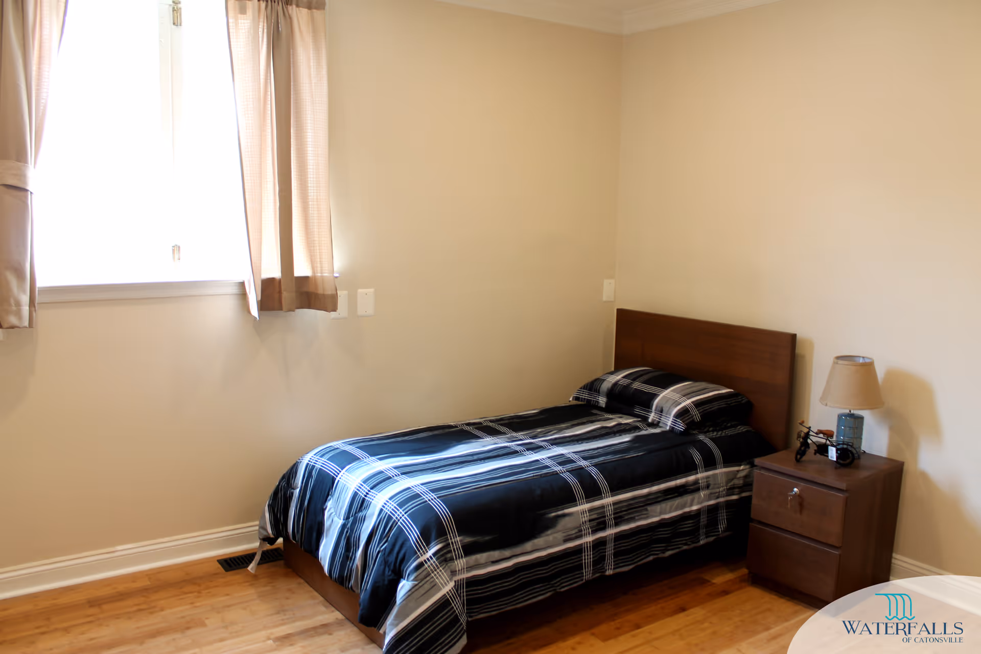 Sparsely furnished bedroom with a single bed in black plaid bedding, a wooden nightstand and lamp beside a curtained window.