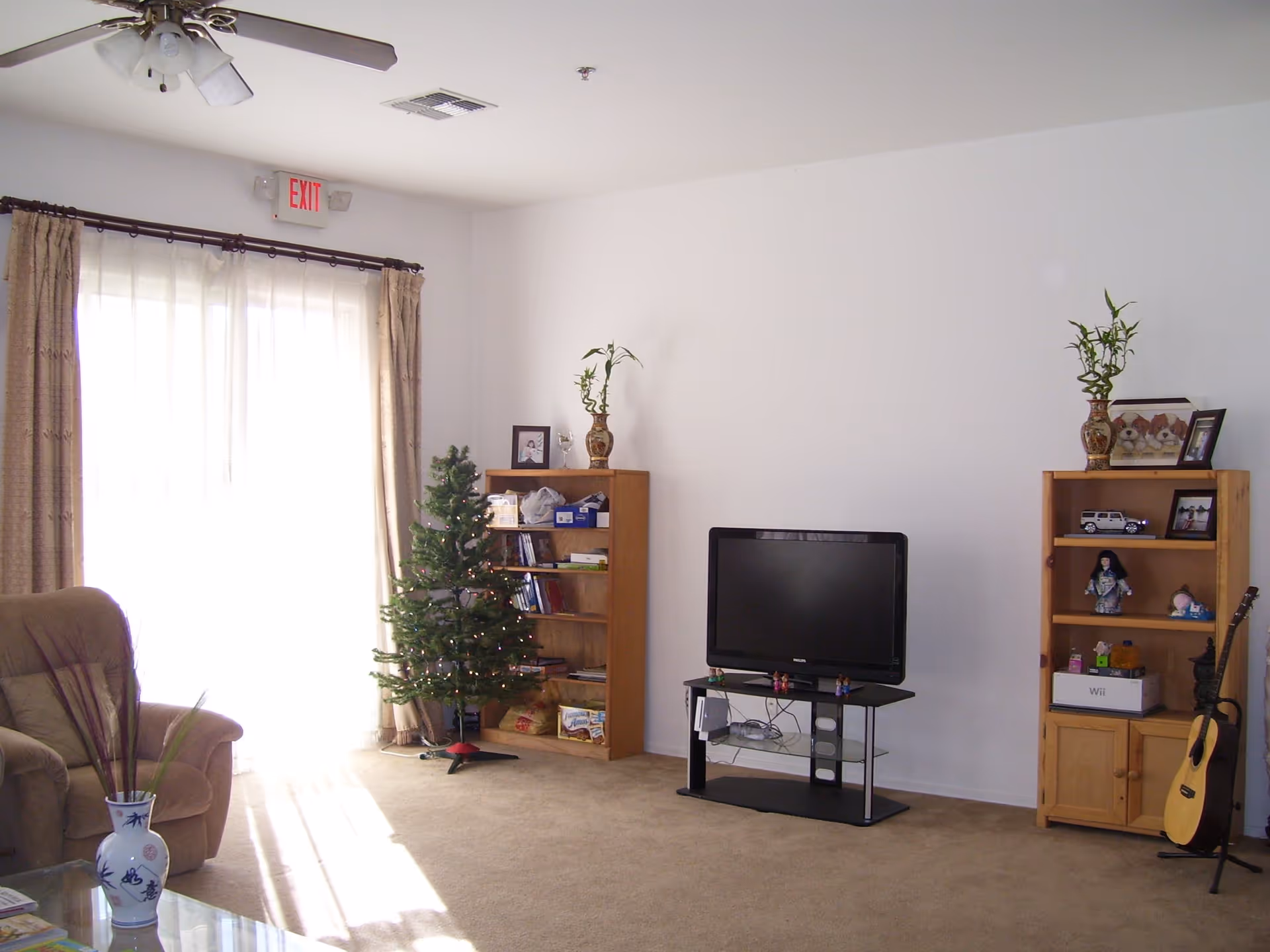 Bright living room with a TV on a stand between two wooden bookshelves, a small decorated Christmas tree, an armchair, and a sliding glass door with curtains.