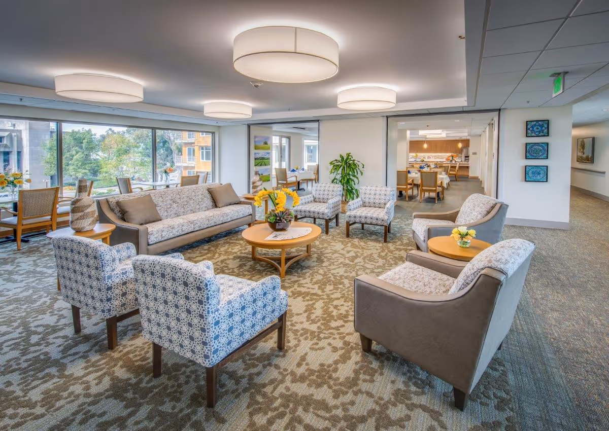 A spacious and well-lit living room area in a senior living facility with patterned armchairs and a sofa arranged around a round wooden coffee table with a flower arrangement. Large windows provide natural light and views of trees and buildings outside. The room features modern ceiling lights, carpeted floors, and additional seating and dining areas visible in the background.