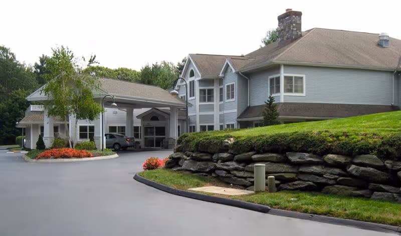 Exterior view of a senior living facility building with gray siding and a stone chimney. The building has multiple windows and a covered entrance with a driveway. There are landscaped areas with green grass, a rock wall, and flower beds with red flowers near the entrance.