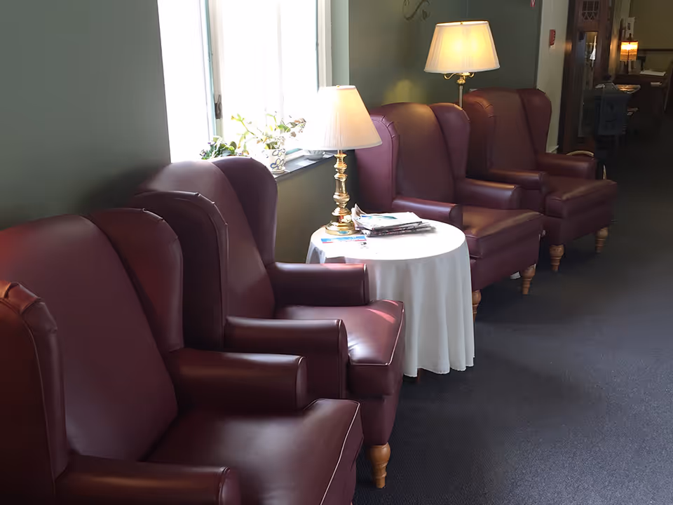 A row of four maroon leather armchairs lined up against a wall with two table lamps on a round table covered with a white tablecloth. There is a window with a small plant on the windowsill, and some magazines on the table. The setting appears to be a quiet indoor common area or lounge.