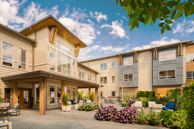 Sunlit courtyard with seating, flowering planters, and a multi-story assisted living building under a blue sky.