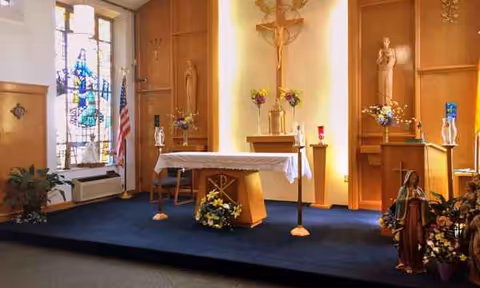 Interior chapel in a senior facility with altar, crucifix, statues, flag, flowers and stained glass.