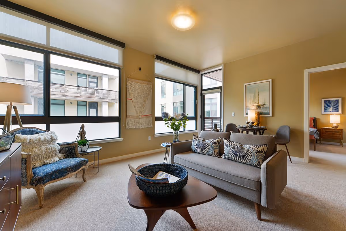Bright furnished living room with a gray sofa, patterned pillows, an armchair, coffee table and large windows overlooking a courtyard.
