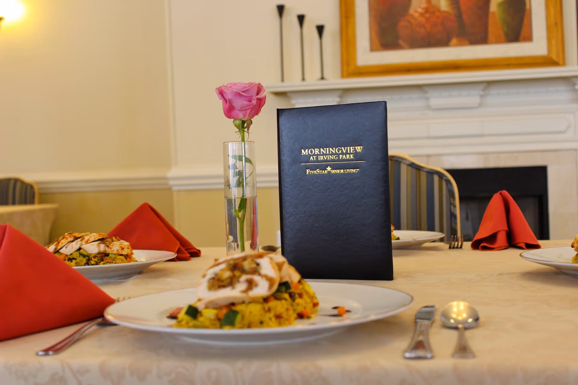 A dining table set with plates of food, red folded napkins, silverware, and a single pink rose in a glass vase. In the center of the table is a black menu with gold text that reads 'Morningview at Irving Park Five Star Senior Living'. The background shows a fireplace and a framed painting above it.