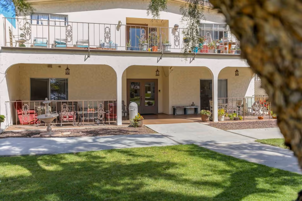 Front façade of a two-story assisted living building with balconies, arched ground-floor entrances, outdoor seating and a lawn.