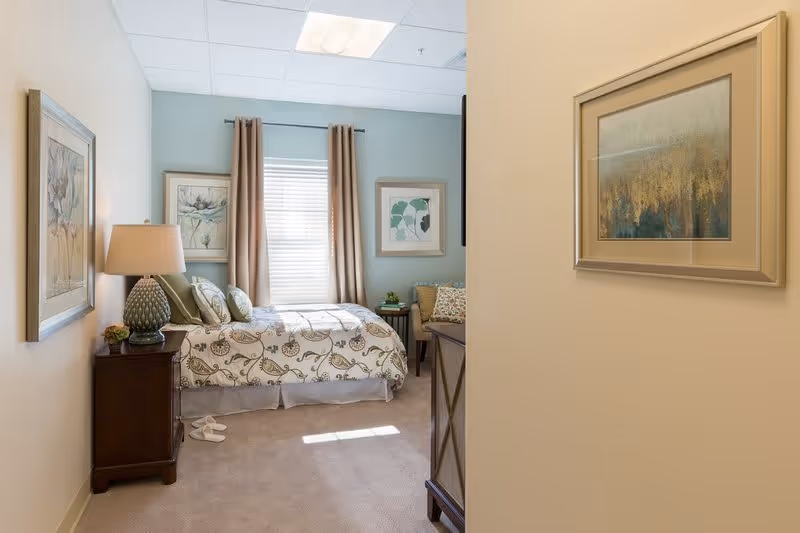 A cozy bedroom with a bed covered in a floral patterned bedspread, two framed pictures on the walls, a wooden nightstand with a decorative lamp and small plant, beige curtains on the window, and a pair of white slippers on the carpeted floor.