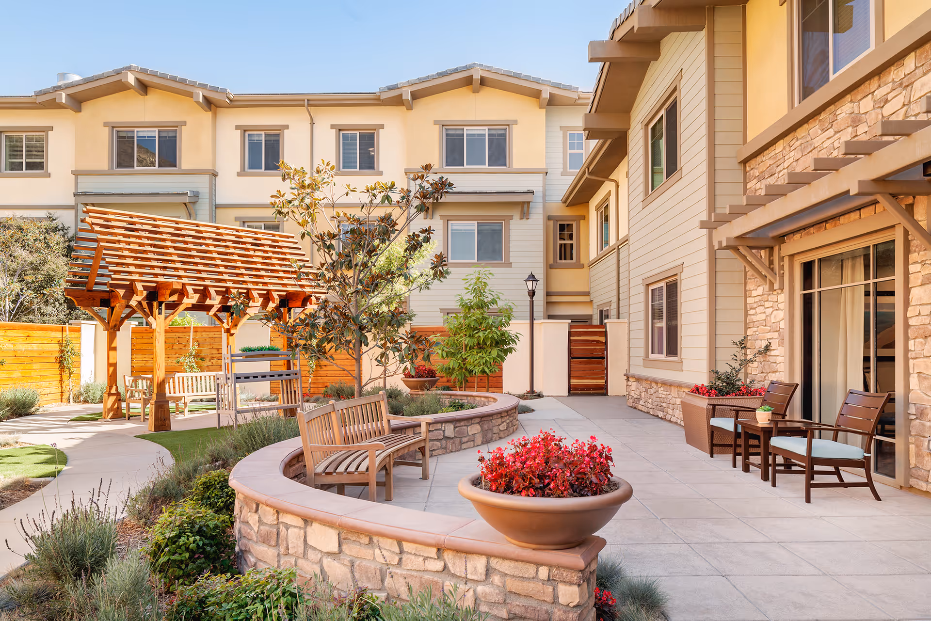 Outdoor courtyard area of a senior living facility with wooden benches, a large planter with red flowers, a wooden pergola, and a paved walkway surrounded by greenery. The building exterior features beige siding and stone accents with windows and patio doors.