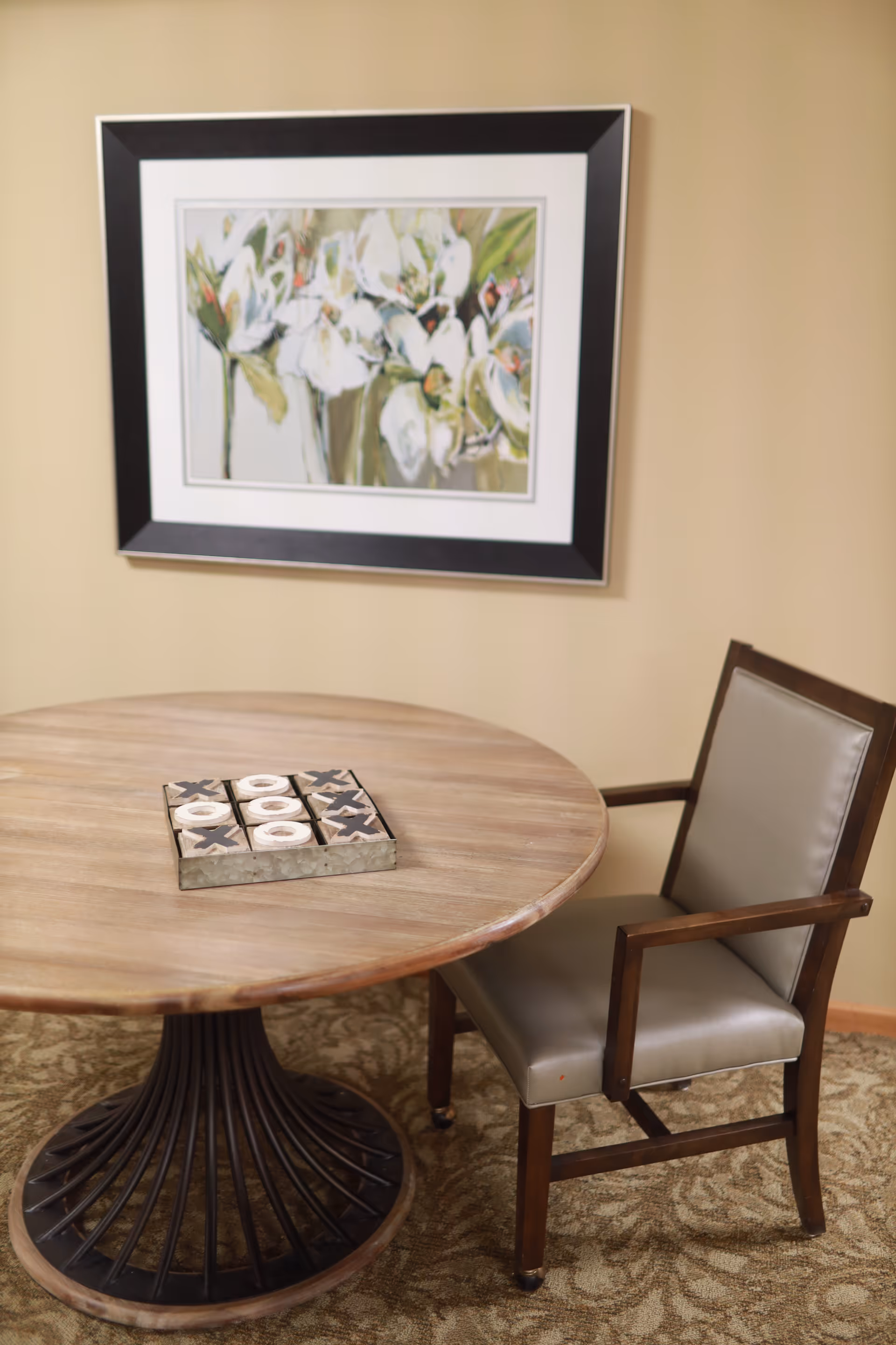 A round wooden table with a tic-tac-toe game set on top, next to a cushioned wooden chair. A framed painting of white flowers hangs on the beige wall behind the table.