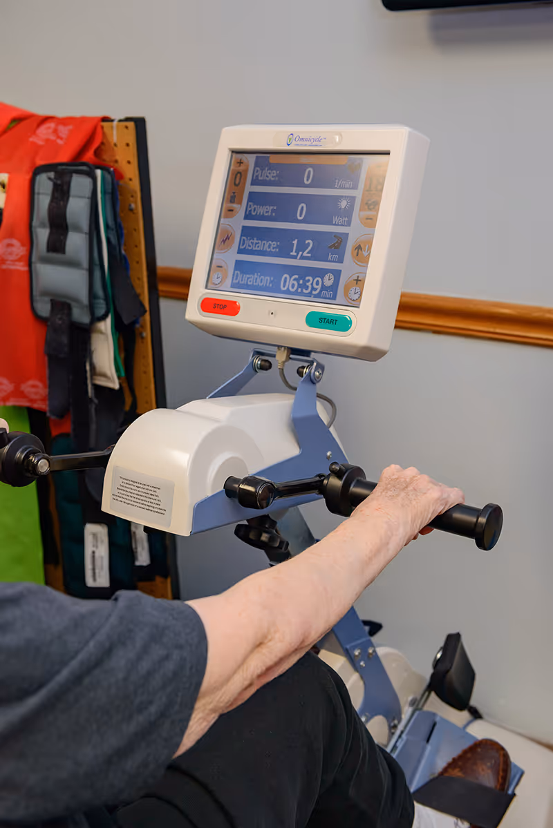 Close-up of an elderly person's arm using a hand cycle exercise machine with a digital display showing pulse, power, distance, and duration. Colorful exercise weights or equipment are visible in the background.