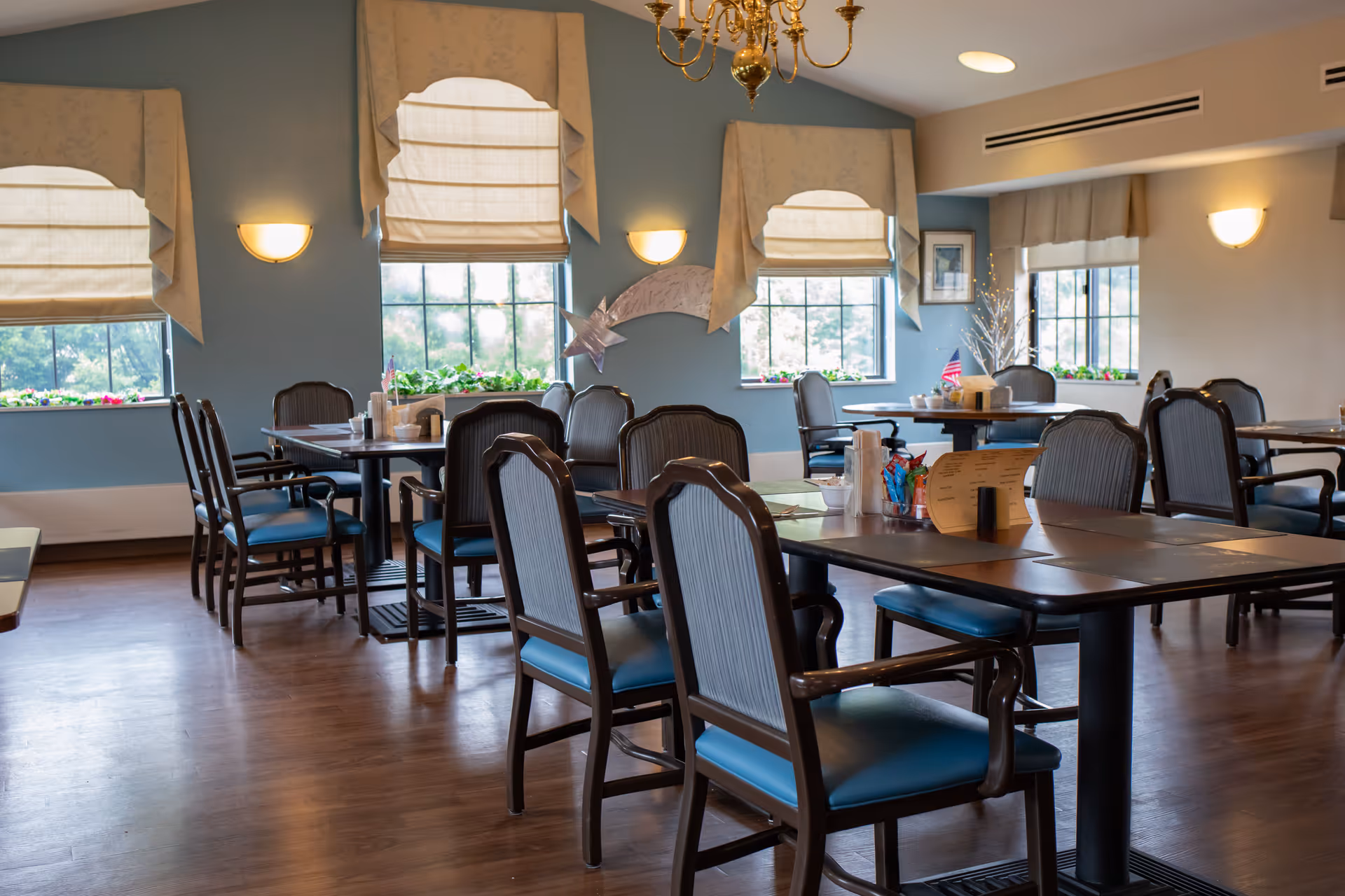 A dining room with several tables and chairs arranged neatly. The room has large windows with beige curtains and valances, letting in natural light. The walls are painted blue and beige, and there are wall-mounted lights and a chandelier hanging from the ceiling. Some tables have menus, condiments, and small American flags on them.
