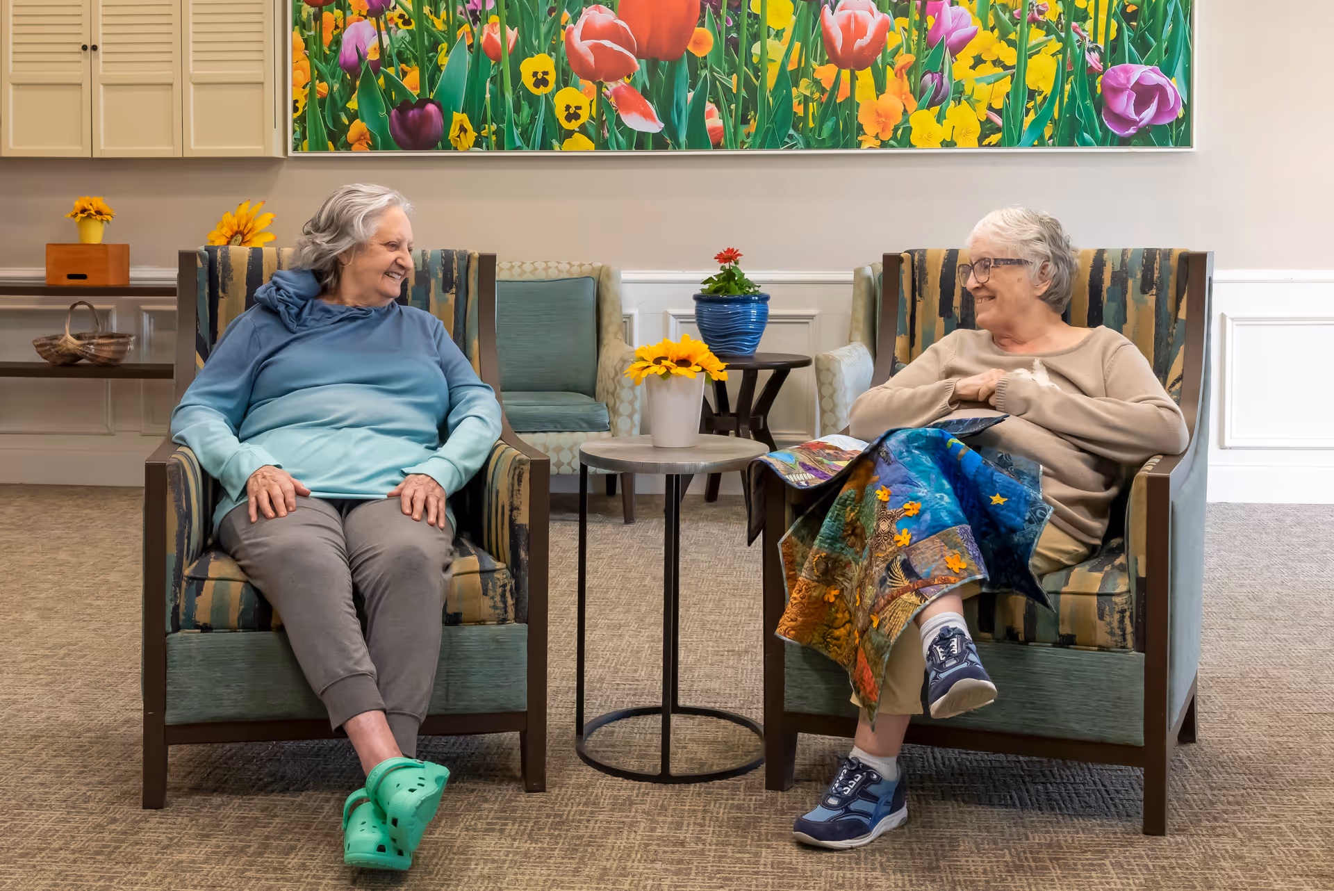 Two elderly women sitting in armchairs facing each other and smiling in a cozy room with a colorful floral painting on the wall behind them. A small round table with a potted plant is placed between the chairs.