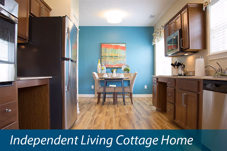 A bright kitchen and dining area with wooden cabinets, stainless steel appliances including a refrigerator and microwave, a small dining table with two chairs, and a blue accent wall with colorful artwork. The floor is wood, and there are windows letting in natural light.