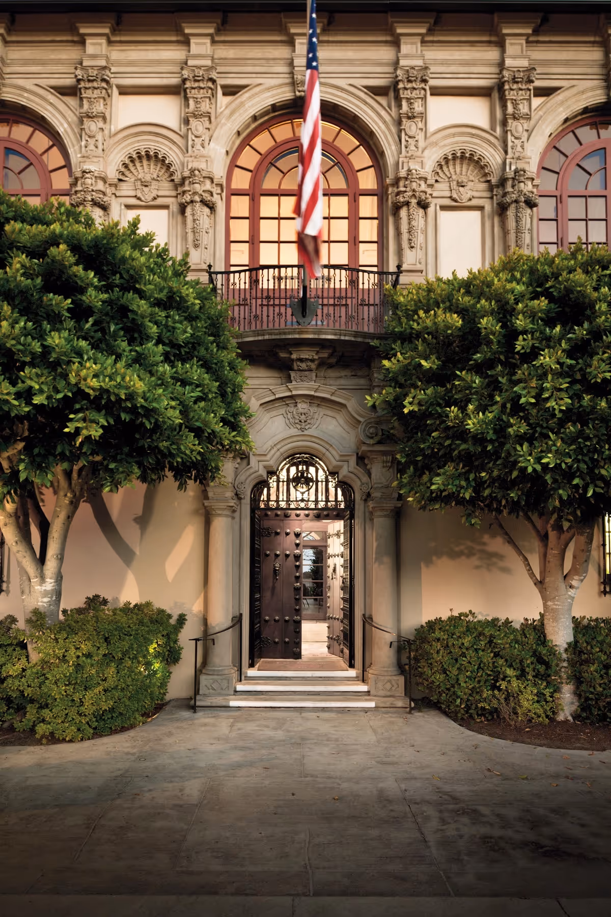 Ornate historic building entrance with an open studded wooden door, flanked by trimmed trees and topped by an American flag.