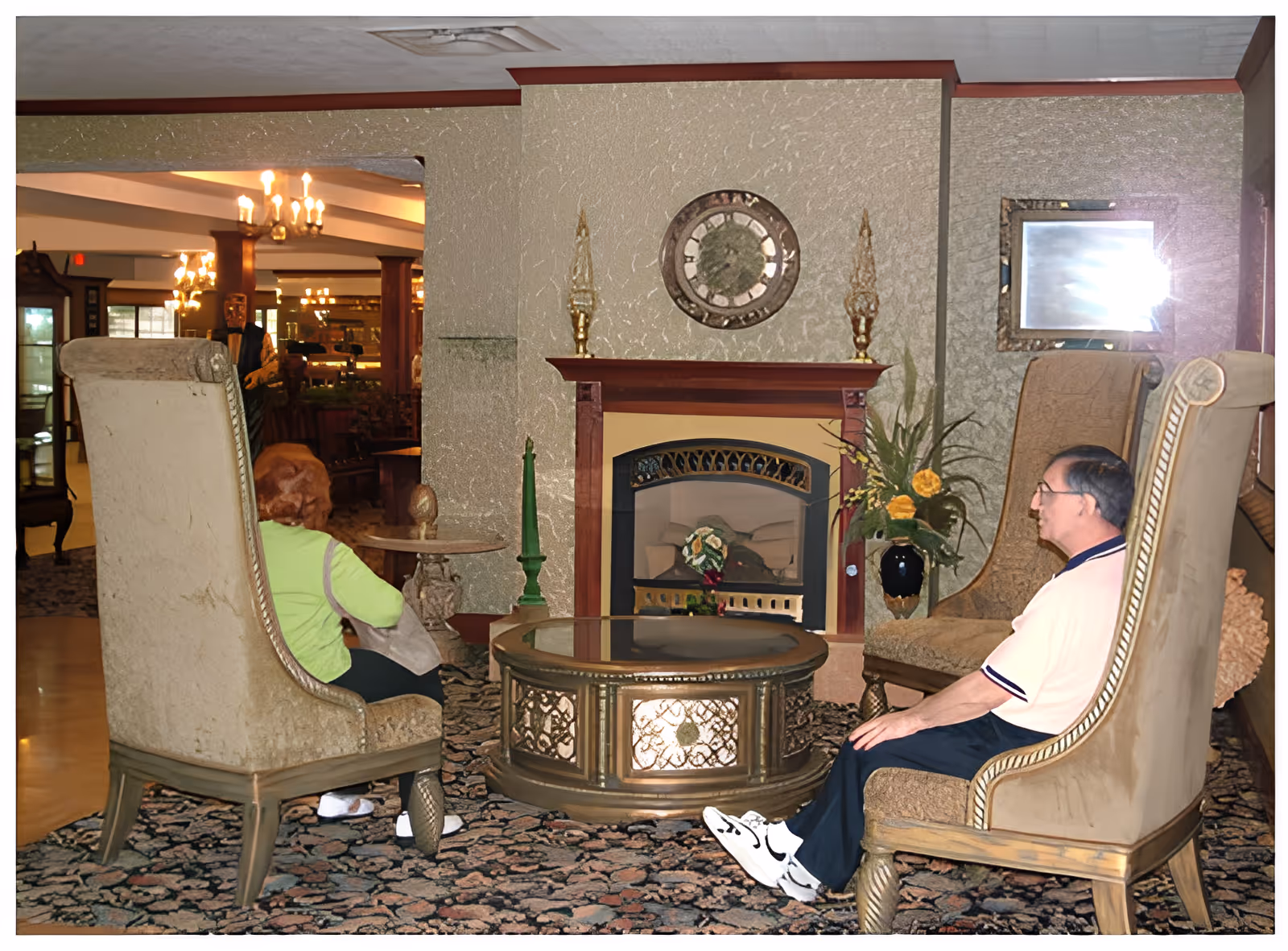Two elderly individuals sitting in high-backed armchairs facing each other in a cozy lounge area with a decorative fireplace, a round glass-top coffee table, and floral arrangements. The room has patterned carpet and warm lighting from chandeliers in the background.