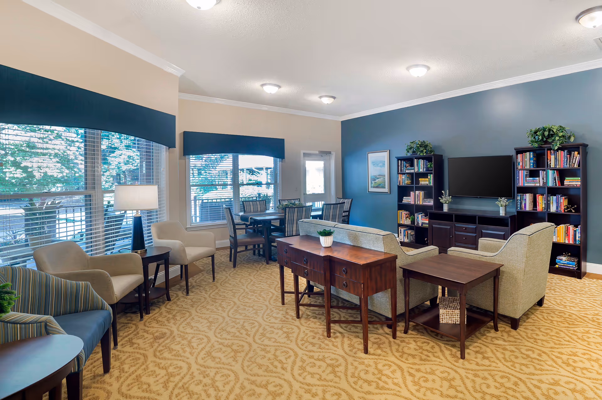 A bright and spacious living room area with large windows covered by blue valances, beige walls, and a blue accent wall. The room features a seating area with a sofa, armchairs, wooden side tables, and a wooden console table with a small plant. There are two dark wood bookshelves filled with books flanking a flat-screen TV mounted on the blue accent wall. A dining table with chairs is positioned near the windows, and the floor is covered with a patterned beige carpet.
