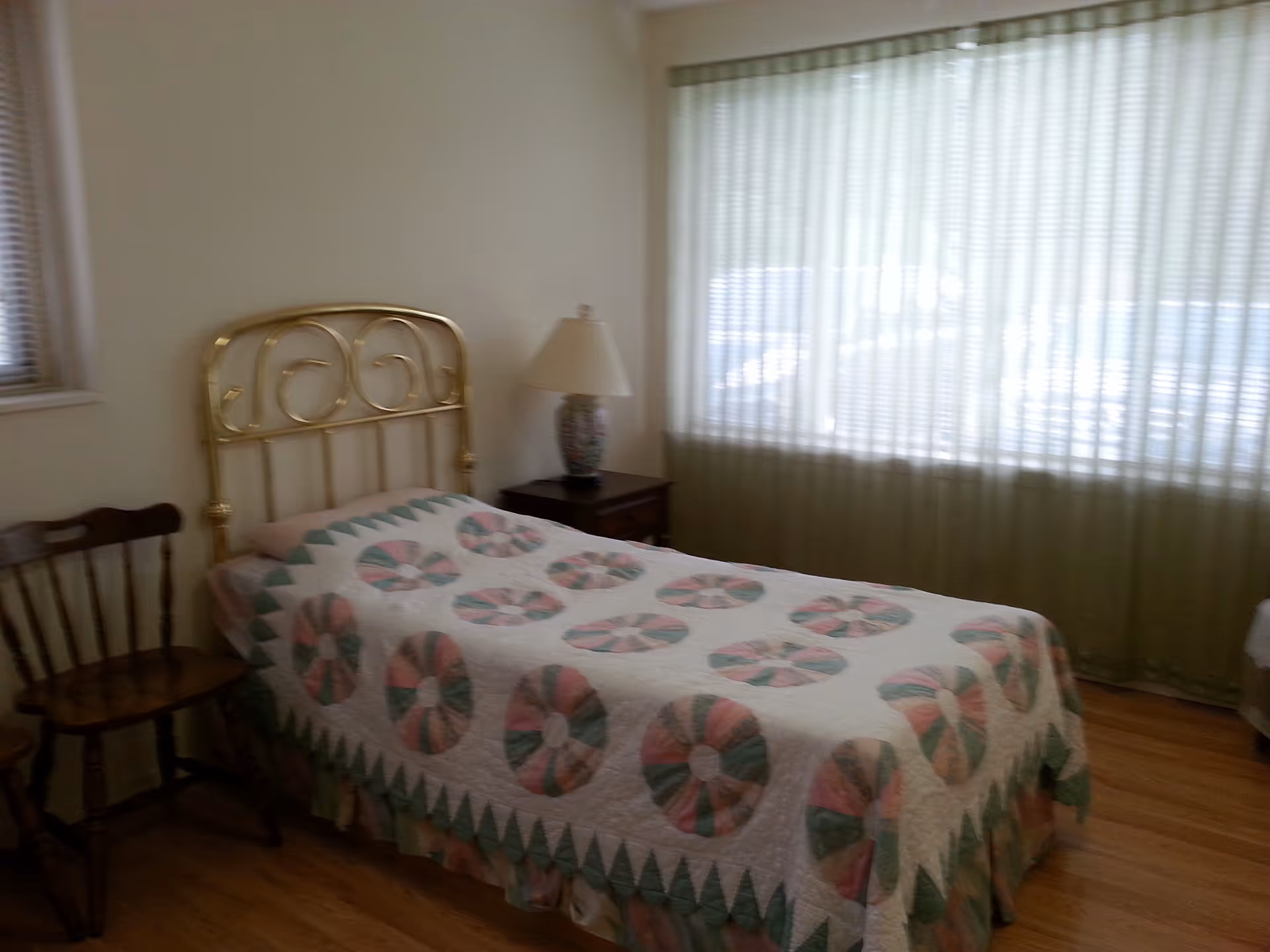 Small bedroom with a single bed covered by a patterned quilt, a brass headboard, nightstand with a lamp, wooden chairs, and a large curtained window.