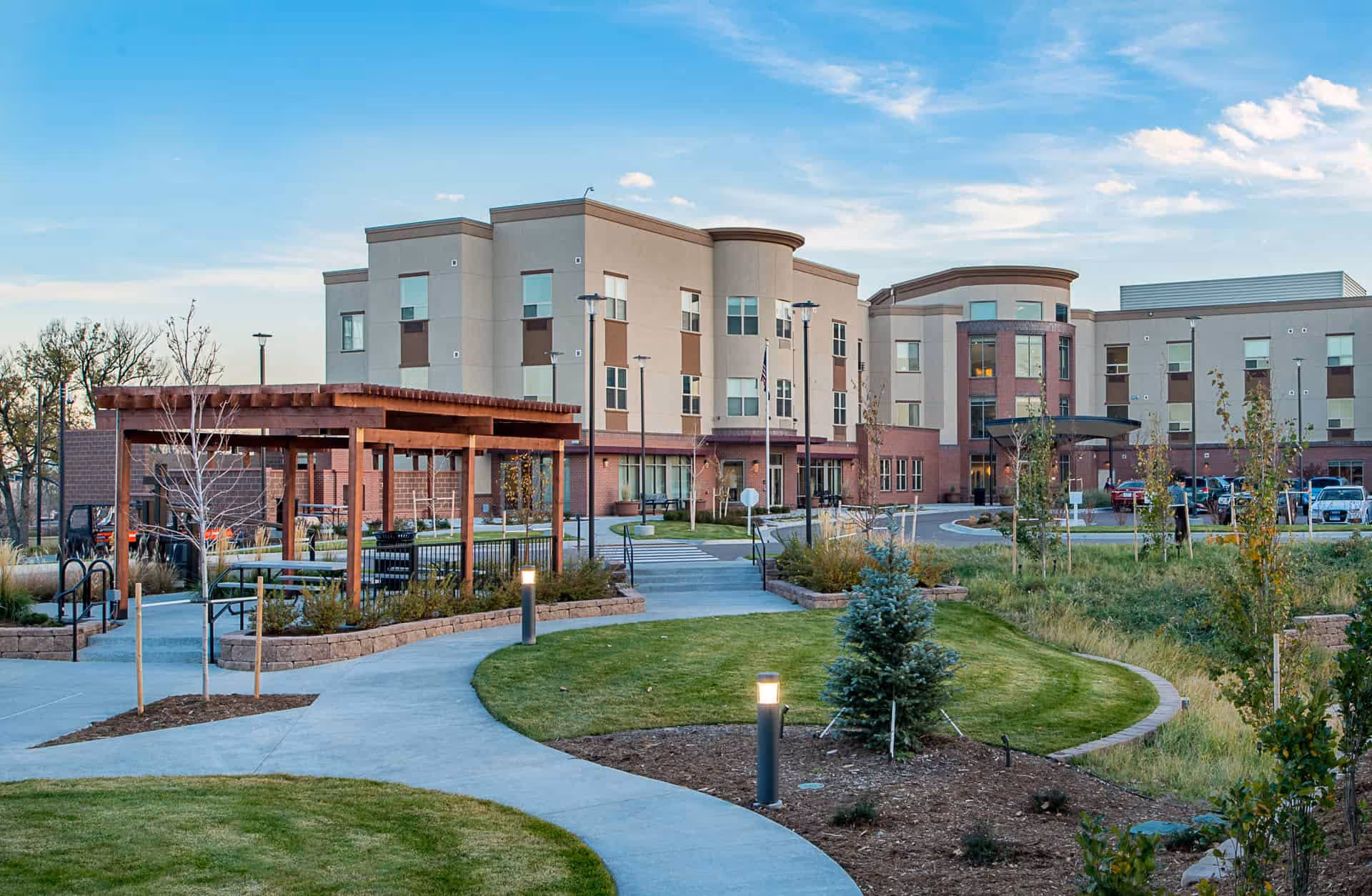 Exterior view of the Village at Belmar senior living facility showing a modern three-story building with a curved entrance, surrounded by landscaped gardens, a paved walkway, small trees, and a wooden pergola with benches underneath. The sky is clear with some clouds.