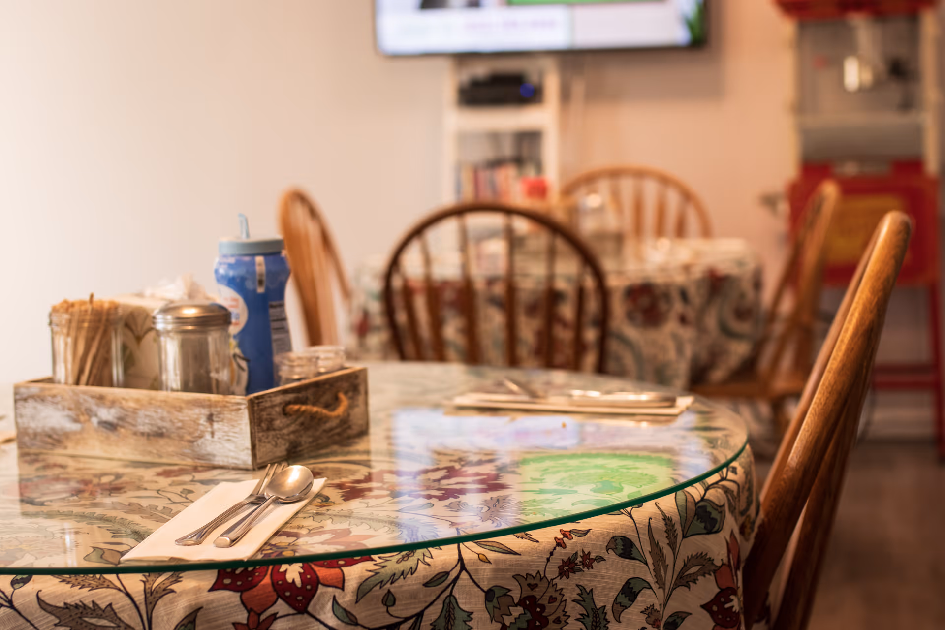 A close-up view of a dining table with a glass top and floral tablecloth, set with a spoon and fork on a napkin. A wooden tray on the table holds condiments and utensils. In the background, there are wooden chairs and another table with a similar setup in a cozy dining area.