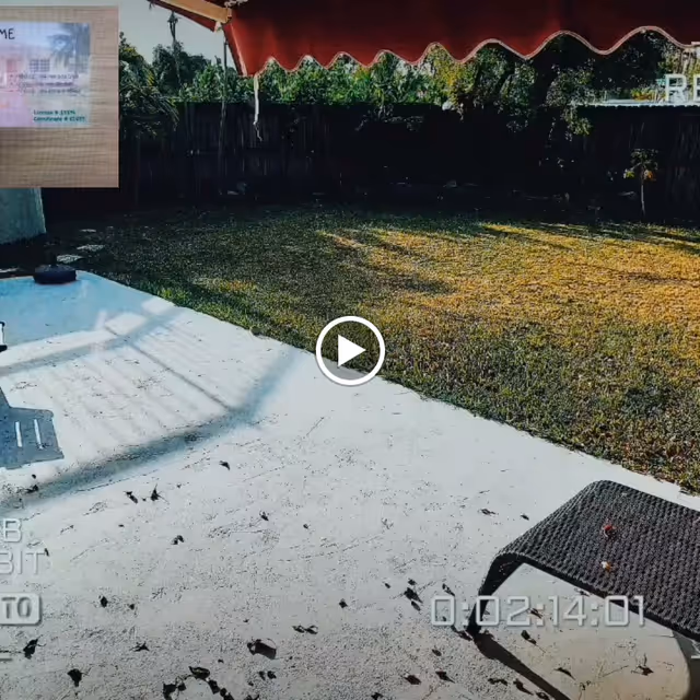 Outdoor patio area with a concrete floor and a lawn in the background, partially shaded by a red awning. There is a black woven chair on the right side and a wooden fence with greenery behind it.