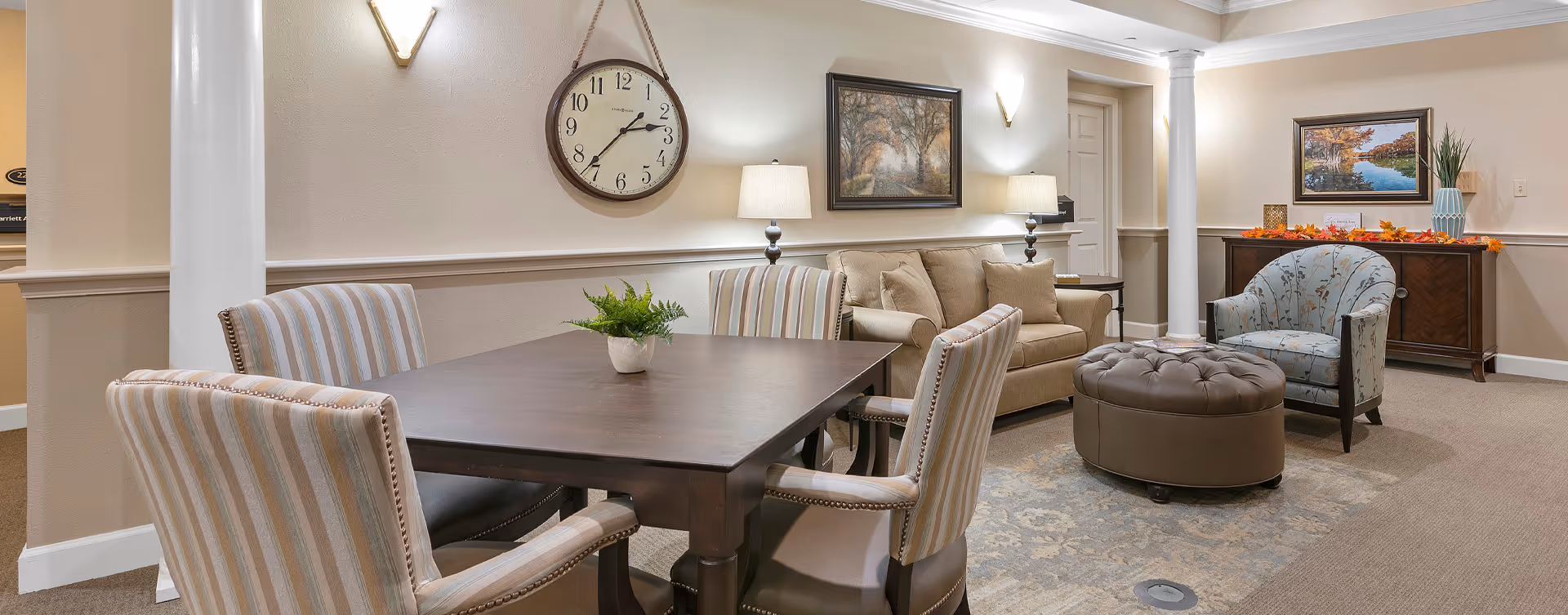 Well-lit communal living area with a dining table and upholstered chairs, a sofa, armchair and ottoman, wall clock and artwork.