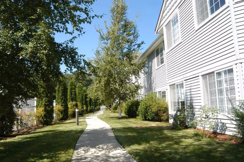 A paved walkway curves through a grassy outdoor area with trees and shrubs on both sides. On the right side, there is a white building with multiple windows. The sky is clear and blue.