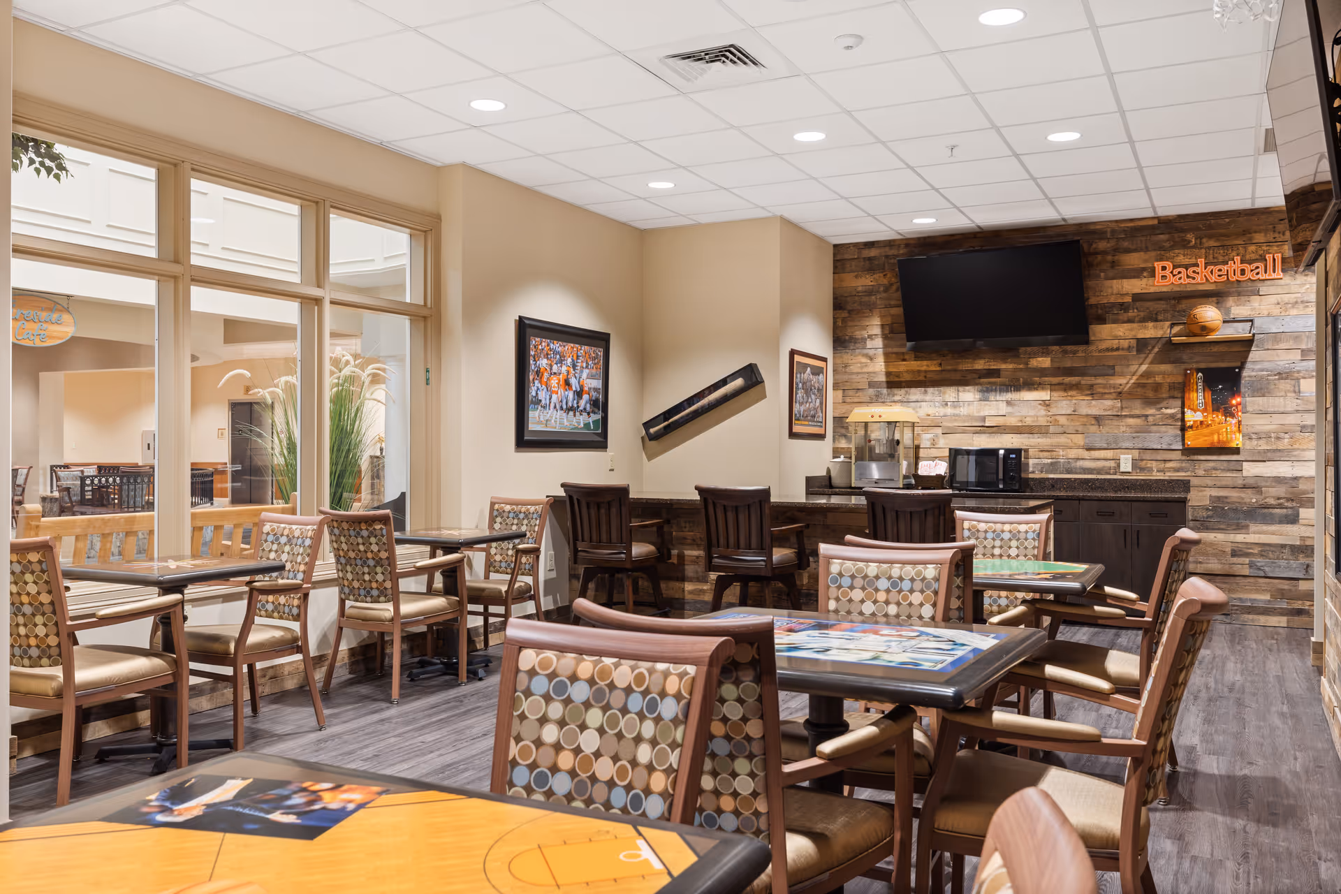 A cozy dining area with several tables and chairs featuring patterned upholstery. The room has large windows letting in natural light, a wooden accent wall with a mounted TV, a popcorn machine, microwave, and basketball-themed decor including a basketball and framed pictures.