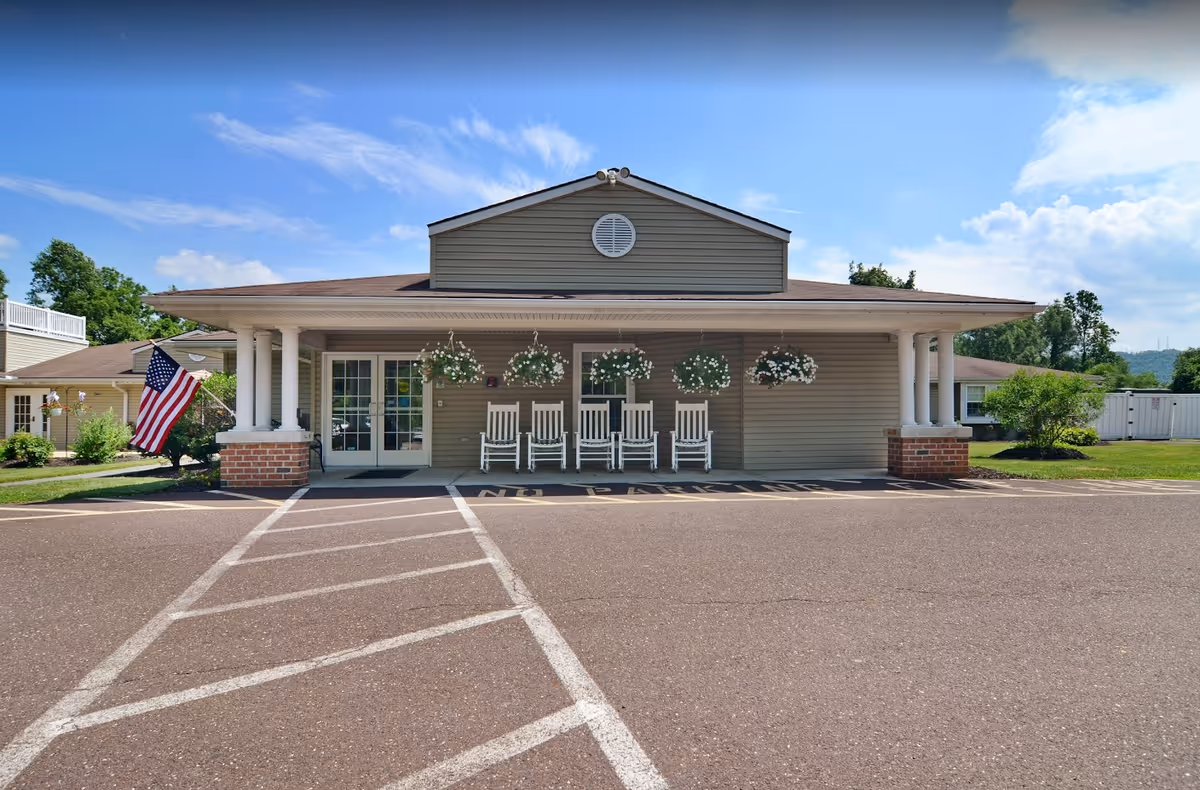 Front exterior view of a single-story building with a covered porch featuring four white rocking chairs and hanging flower baskets. An American flag is displayed on the left side near the entrance. The building is surrounded by greenery and a clear blue sky with some clouds.