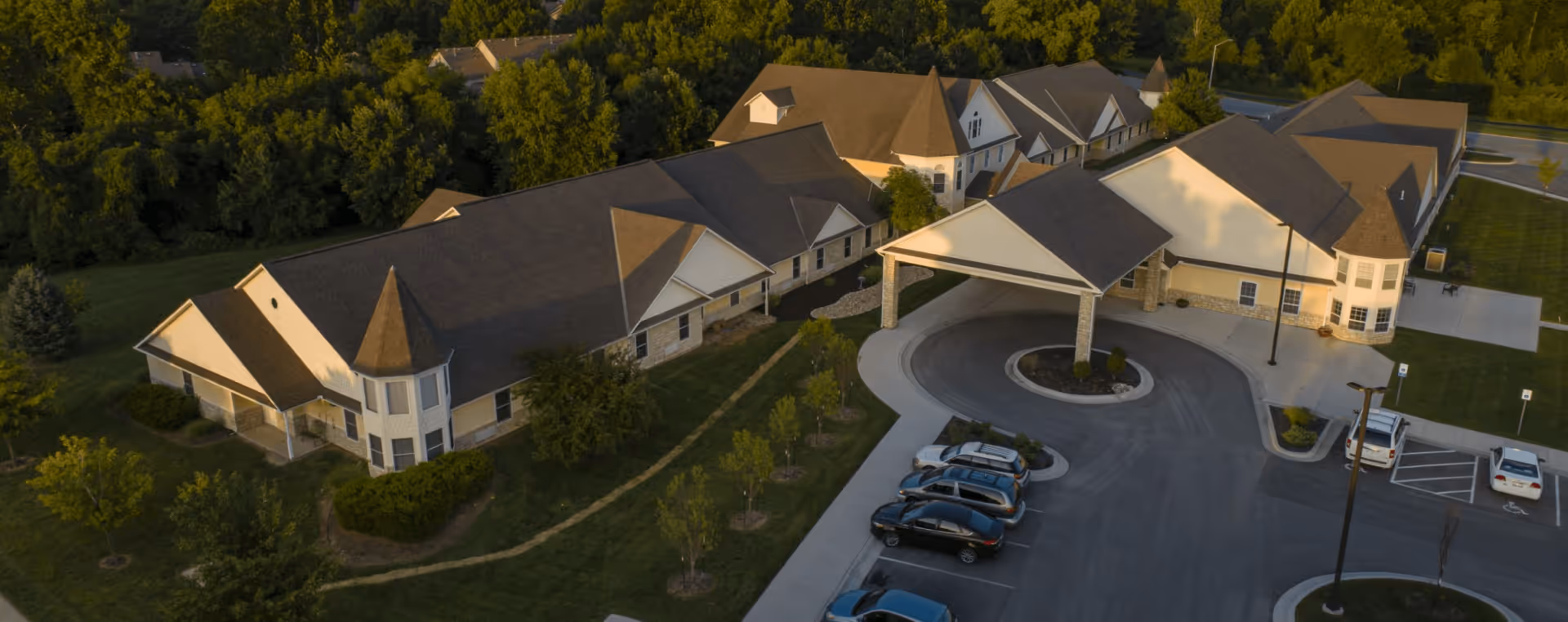 Aerial view of Homestead Assisted Living & Memory Care of Overland Park showing a large building with multiple peaked roofs surrounded by trees and greenery. There is a circular driveway with a covered entrance and several parked cars in the parking lot.