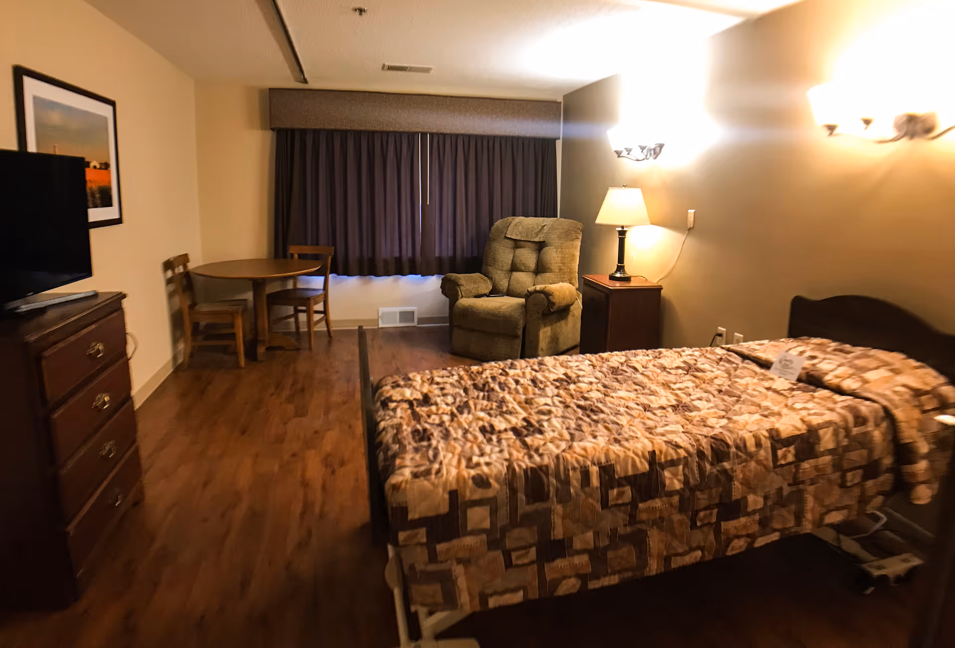 Furnished senior bedroom with a single bed, recliner, small round table and chairs, dresser with TV, and bedside lamp.