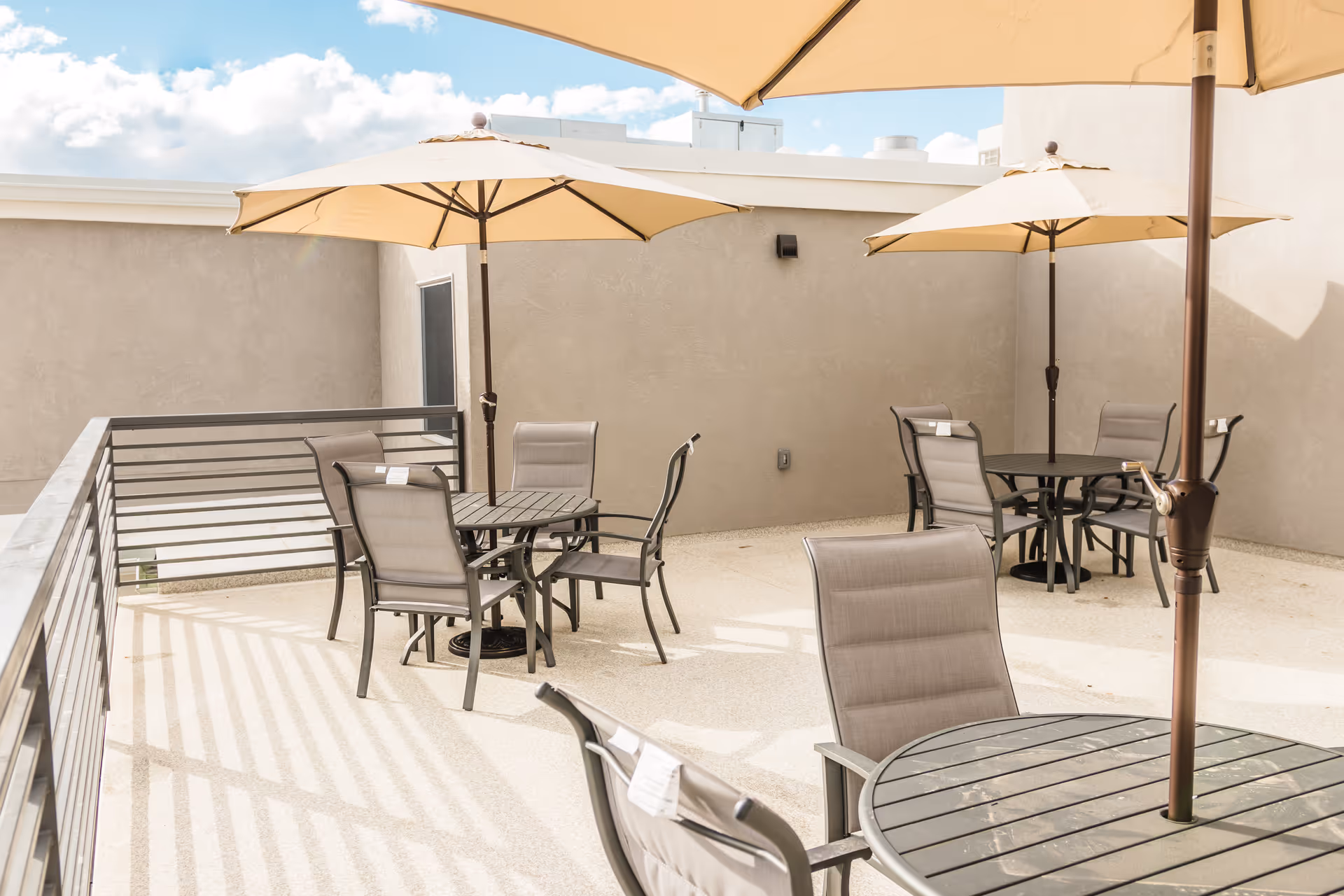 Outdoor patio area with multiple round tables and chairs, each table shaded by a large beige umbrella. The space is enclosed by beige walls and a metal railing, under a partly cloudy blue sky.