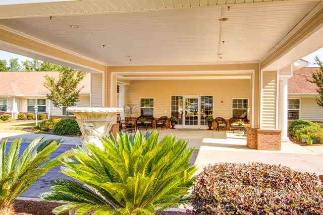 Covered entrance area of a senior living facility with outdoor seating including chairs and small tables. There are green plants and shrubs in the foreground, and the building has beige siding with white trim and a red roof.