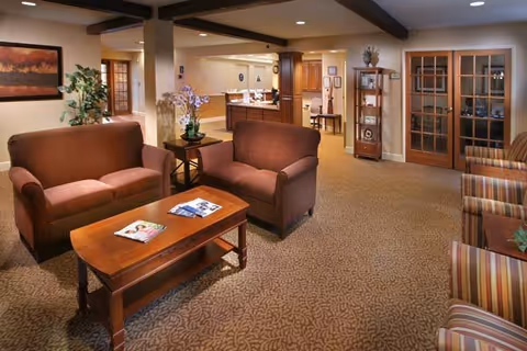 A cozy senior living facility common area with two brown upholstered loveseats facing a wooden coffee table with magazines on it. The room has beige patterned carpet, a wooden side table with a potted orchid, a wooden display cabinet, and French doors with glass panes. The walls are decorated with framed artwork and the ceiling has exposed wooden beams.